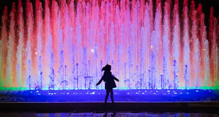 Silueta de una chica bailando frente a una fuente multicolor iluminada de noche.