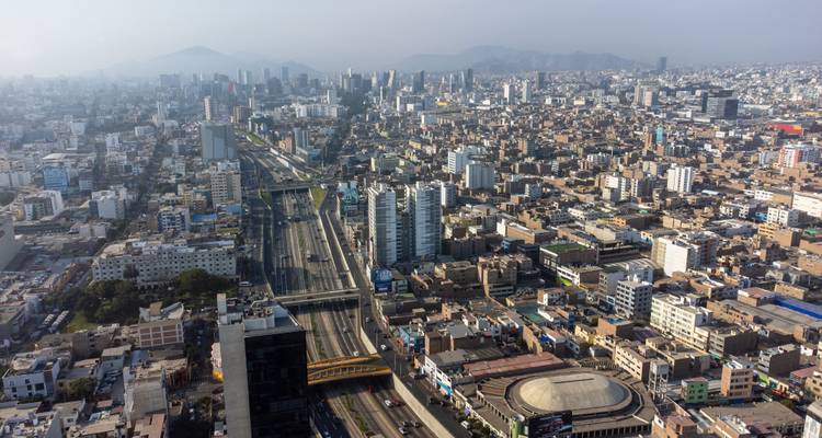Amplio paisaje urbano de Lima con edificios de gran altura, autopista y montañas lejanas y brumosas bajo la luz del mediodía.