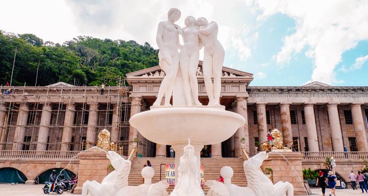 Une fontaine avec statue en marbre blanc s'élève devant un bâtiment néoclassique adossé à une colline boisée sous un ciel dégagé.