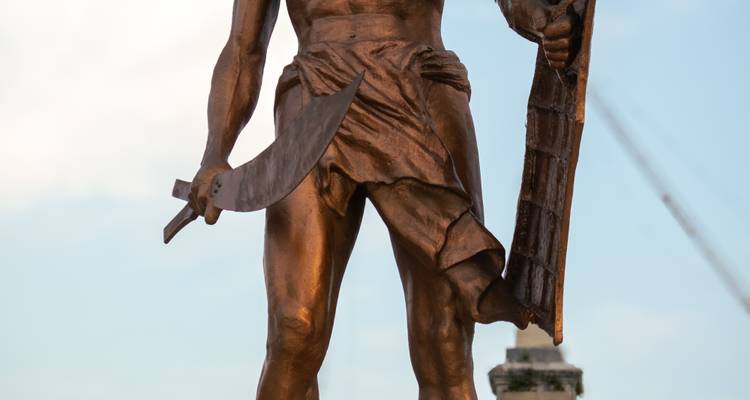 Gros plan d'une statue de guerrier en bronze tenant une lame courbe sur un fond de ciel pâle.