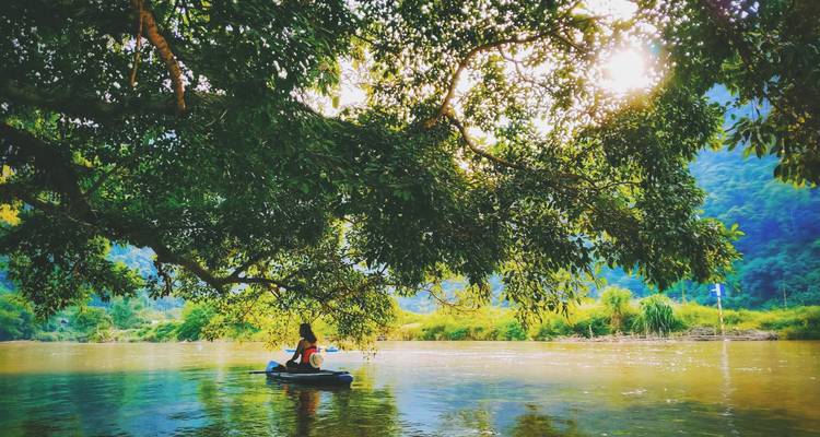 Un remero solitario se desliza bajo ramas frondosas en un río tranquilo y salpicado de sol, bordeado por colinas de selva.