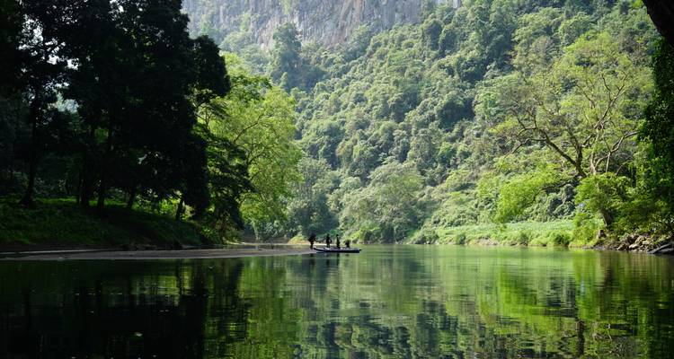 El río tranquilo refleja la densa selva verde y los acantilados mientras un pequeño bote lleva pasajeros a lo lejos.