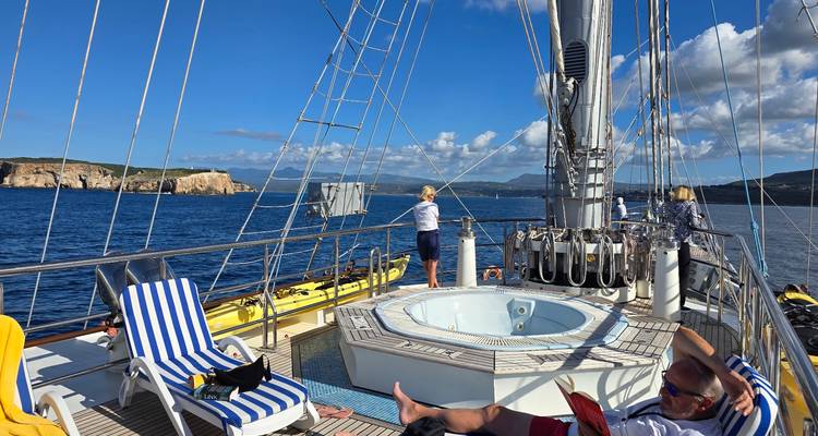 Des passagers se détendent près d'un jacuzzi sur le pont d'un navire de luxe à voiles, avec des îlots grecs visibles au loin.