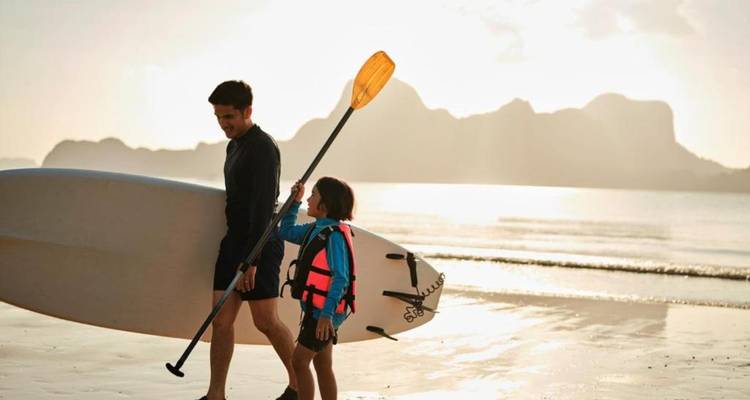 Père et enfant transportant une planche de paddle debout le long d'une plage dorée au lever du soleil
