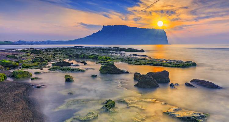 Le soleil se lève sur un plateau d'île volcanique au-delà de rochers côtiers couverts de mousse et d'une mer cristalline.