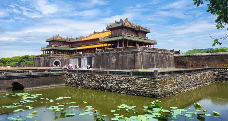 Ciudadela imperial histórica en Hue con puerta ornamentada, foso lleno de lotos y turistas cruzando el puente bajo un cielo azul.