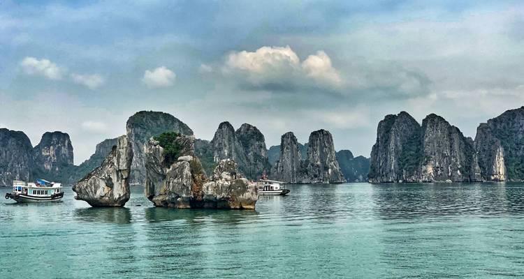 Pilares de piedra caliza dramáticos que se alzan desde aguas color verde azulado tranquilo en la brumosa Bahía de Ha Long con pequeños barcos de crucero.