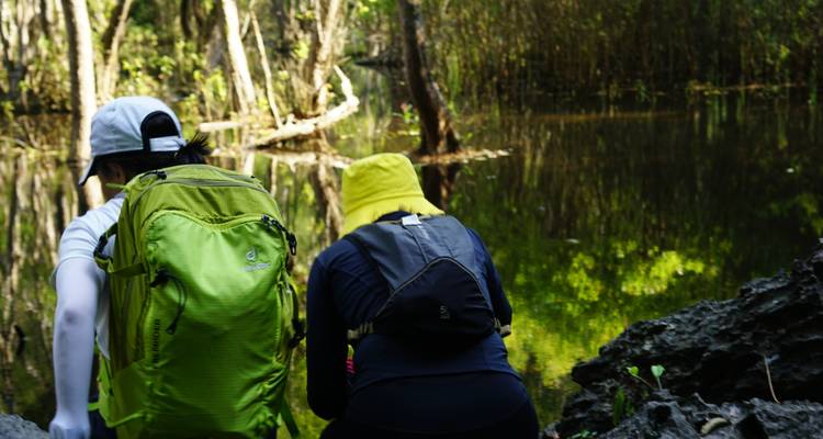 Dos excursionistas con mochilas haciendo una pausa junto a una laguna forestal reflectante.