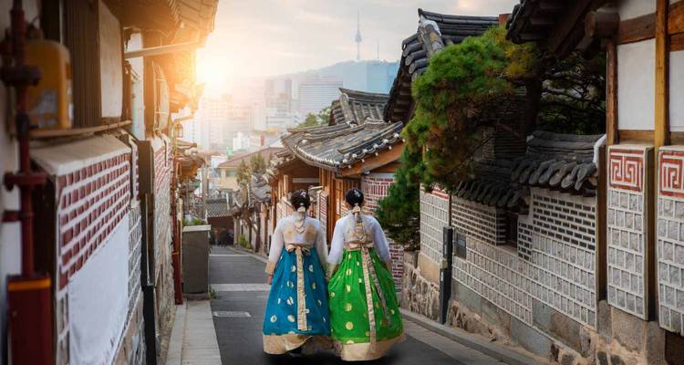 Paire de femmes vêtues de hanbok orné marchant dans une ruelle traditionnelle au coucher du soleil avec la ligne d'horizon de Séoul en arrière-plan.