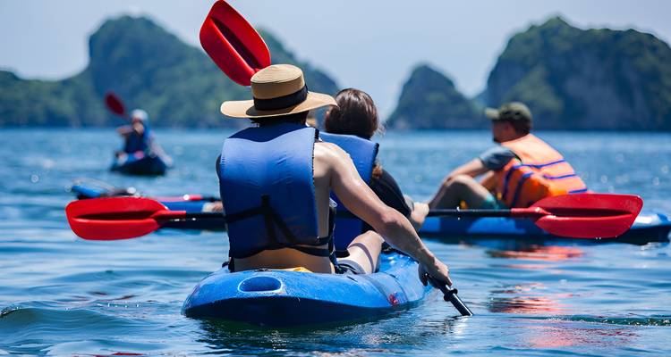 Vista trasera de kayakistas remando hacia formaciones de piedra caliza, fotografiada desde el nivel del agua.