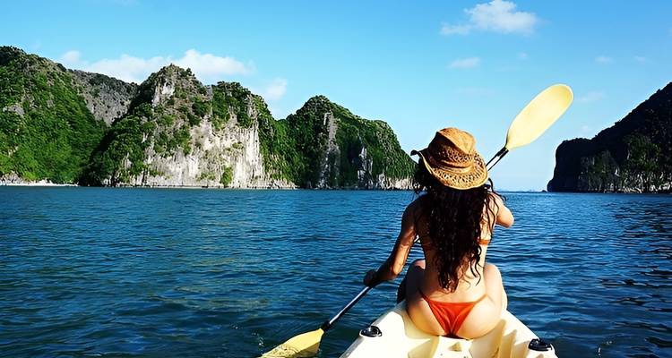 Mujer en traje de baño remando en kayak hacia acantilados de piedra caliza exuberante bajo un cielo azul despejado.