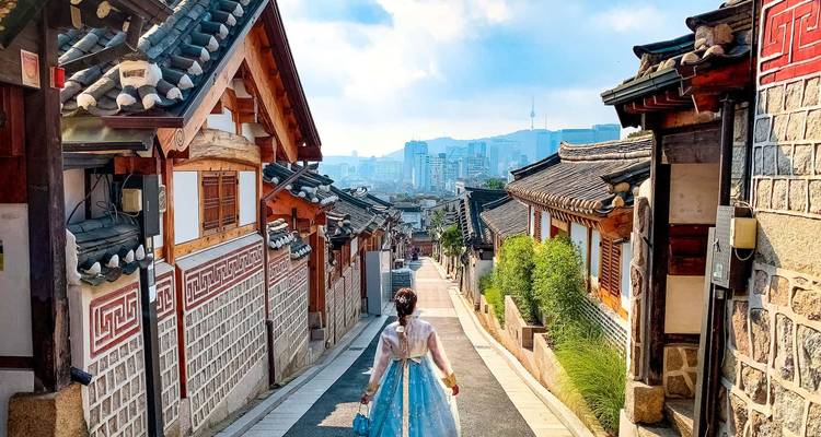Une femme vêtue d'un hanbok traditionnel marchant dans une rue bordée de hanoks en direction de l'horizon de Séoul.