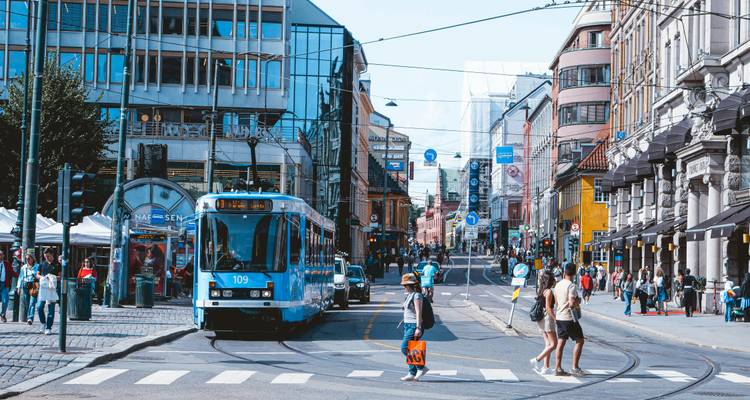Scène animée d'une rue d'Oslo avec un tramway bleu, des piétons traversant et une architecture urbaine variée.