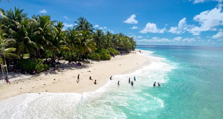 Plage de sable bordée de palmiers où les habitants et les visiteurs s'amusent dans les eaux turquoise peu profondes sous un ciel dégagé.