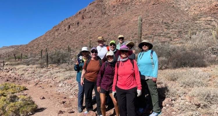 Un groupe de randonneurs pose parmi les arbustes du désert et les grands cactus au pied d'une colline rocheuse.