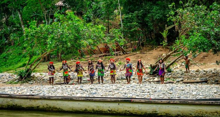 Indigenous villagers in colorful attire stand hand-in-hand on a pebbled riverbank in the jungle.