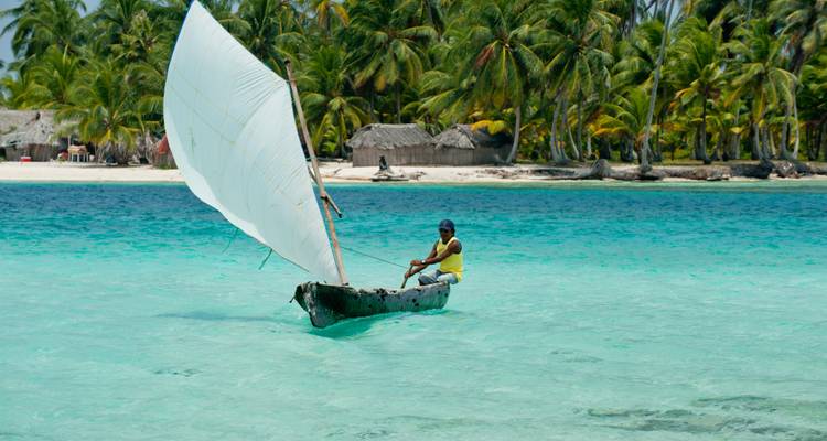 Local sailor navigates a wooden dugout canoe with white sail over clear aqua water near palm islands.