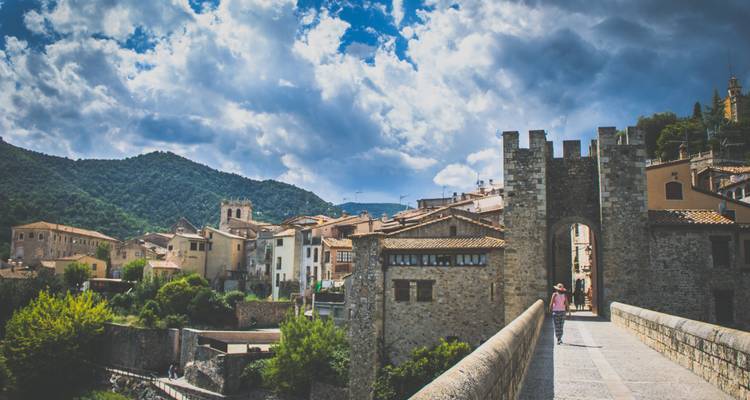 Un pont en pierre mène au village médiéval de Besalú, entouré de collines boisées et de nuages dramatiques.