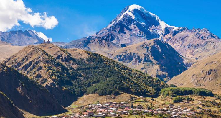 Le Mont Kazbek aux sommets enneigés domine le village de Stepantsminda et les collines ondulantes environnantes sous un ciel bleu dégagé.