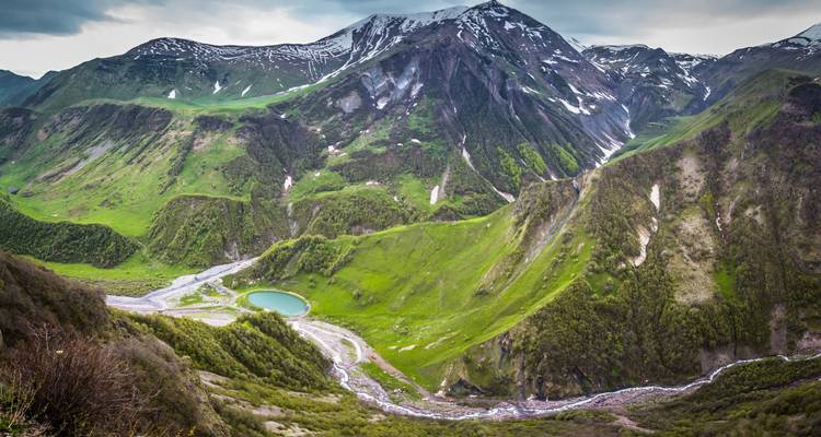 Vue de haut d'une vallée alpine verdoyante avec un petit lac turquoise et des pics enneigés dans le Caucase.