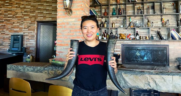 Une femme souriante tient deux cornes à boire traditionnelles à l'intérieur d'un bar à vin géorgien rustique.