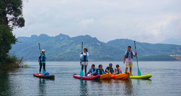 Grupo mixto en kayaks y tablas de paddle flotando en un lago tranquilo rodeado de colinas boscosas