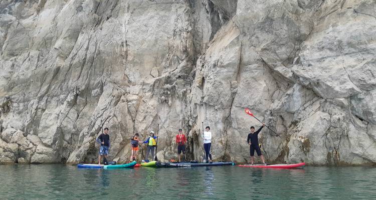 Los practicantes de paddle board descansan apoyados contra una imponente pared de roca gris que se alza directamente desde el agua verde.