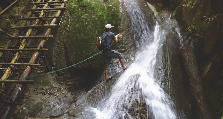 Un homme remonte une cascade étroite en rappel en utilisant des cordes et des échelles en bois dans un canyon de jungle.