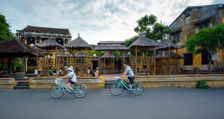 Twee fietsers rijden op vintage fietsen langs rieten bamboe cafés in een erfgoedstraatje.