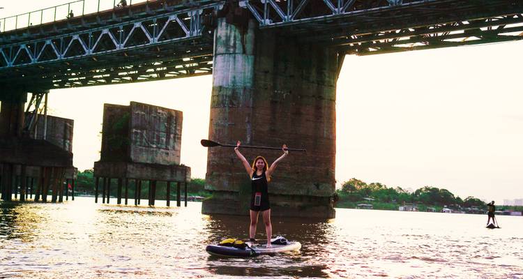 Vrouw die op een paddleboard staat en haar peddel optilt onder een oude stalen brug in de schemering.