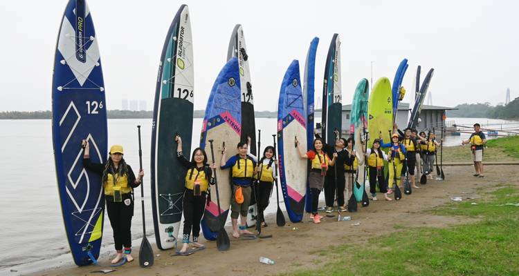 Grote groep die poseert met rechtopstaande paddleboards langs een zandige rivieroever.