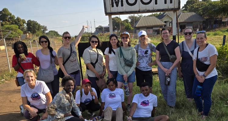 A large tour group stands beside a roadside sign reading "Moshi" for a commemorative photo.