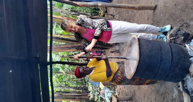 Two women stir a steaming barrel over an open fire in an outdoor kitchen surrounded by banana trees.