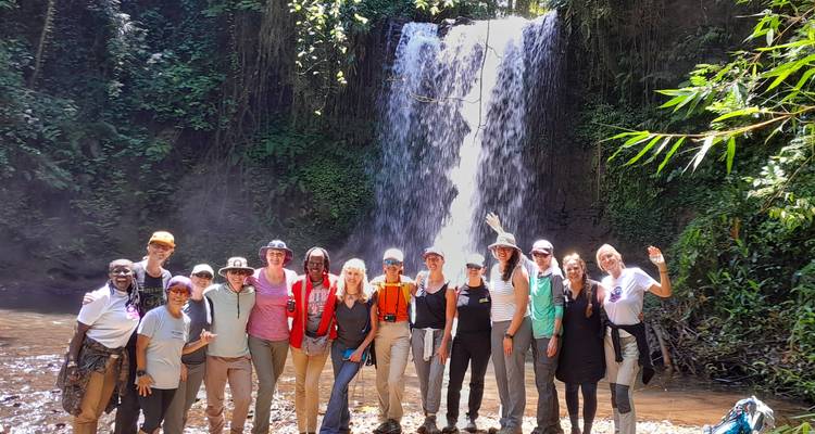 A large tour group poses in front of a waterfall in a shaded jungle setting.