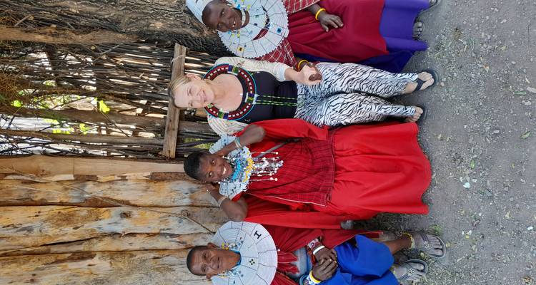 A visitor sits with three Maasai women in traditional red beaded attire outside a wooden fence.