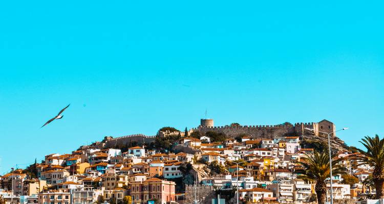 La lumière éclatante du soleil baigne la forteresse perchée sur la colline et les toits en terre cuite de Kavala sous un ciel bleu cyan dégagé.