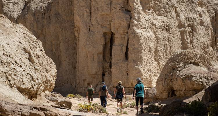 Hikers walk into a narrow, rugged rock canyon under bright desert light.