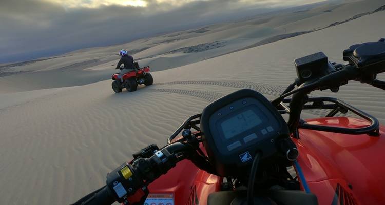 Two riders on red ATVs carve tracks across vast rolling sand dunes under a cloudy sky.
