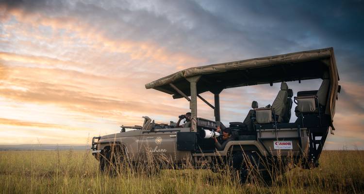Un camión de safari estacionado en la sabana dorada al atardecer bajo un cielo lleno de nubes de colores.