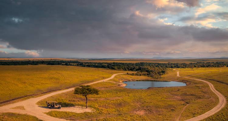 Paisaje aéreo de una vasta sabana con caminos de tierra sinuosos que conducen a un pequeño abrevadero bajo un cielo amenazante.