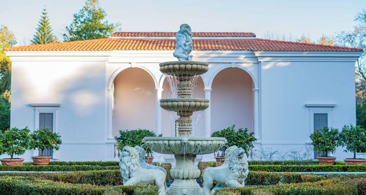 Pabellón clásico de color blanco frente a una fuente de piedra escalonada adornada con estatuas de leones en un jardín bien cuidado.