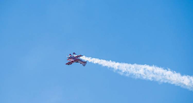 Biplano acrobático solitario subiendo abruptamente con una estela de humo blanco en un cielo azul despejado.