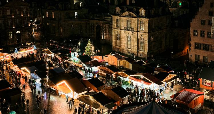 Vue aérienne nocturne d'un marché de Noël illuminé très animé avec des étals en bois et des foules dans une place historique.