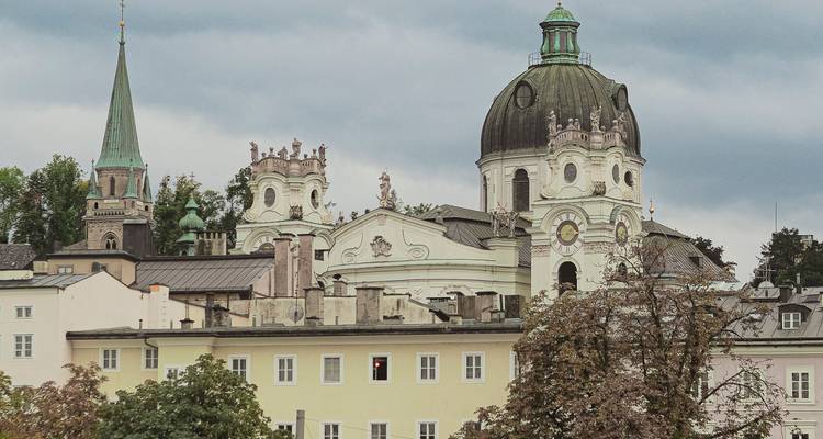 Groupe de dômes et de tours d'églises de Salzbourg s'élevant au-dessus de maisons aux couleurs pastel sous un ciel couvert.