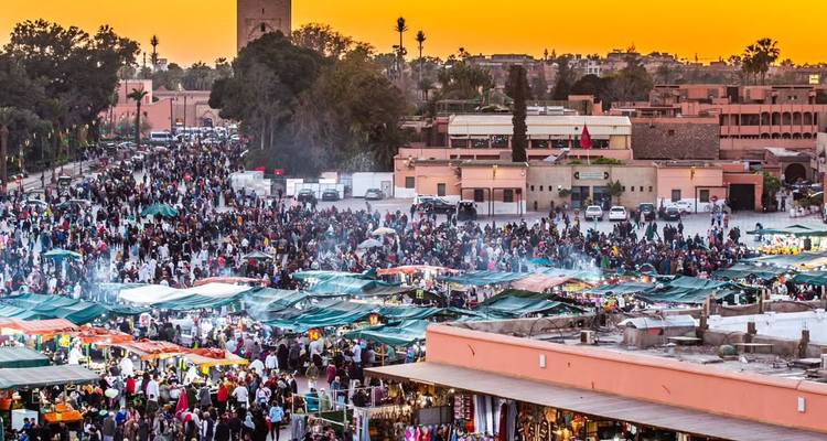 La place Jemaa el-Fnaa animée, remplie d'étals de marché et de foules de personnes sous un ciel au coucher de soleil orange éclatant, avec le minaret de la Koutoubia en silhouette à l'arrière-plan.