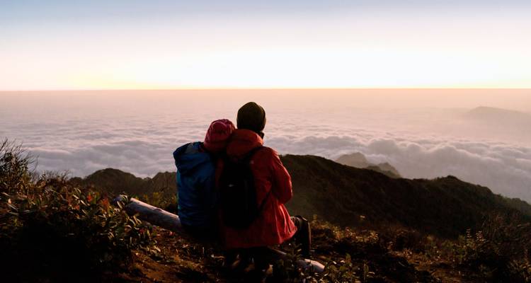 Un couple vêtu de vestes est assis ensemble en observant le lever du soleil au-dessus d'une mer de nuages depuis une crête de montagne