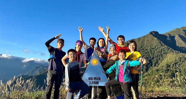 Groupe souriant célébrant au marqueur du sommet indiquant « Nui Muoi 2 225 m » sous un ciel bleu dégagé