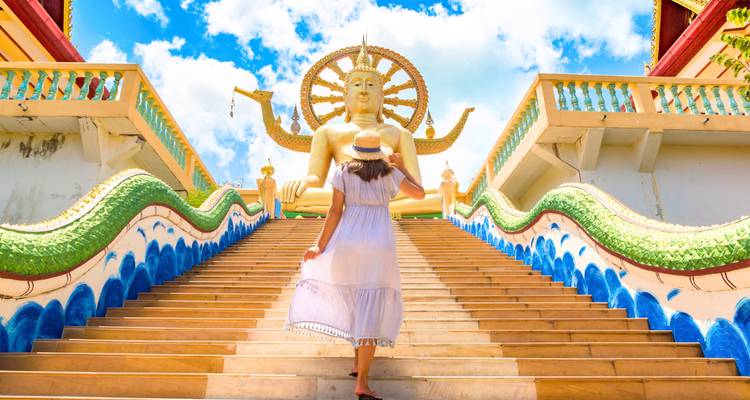 Femme vêtue d'une robe blanche montant les escaliers colorés bordés de serpents vers l'immense statue dorée du Grand Bouddha à Koh Samui.