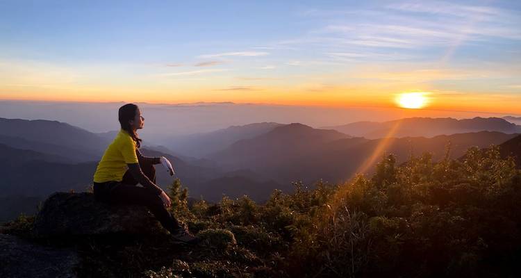 Femme en veste jaune assise sur un rocher regardant un coucher de soleil vibrant sur les chaînes de montagnes en couches