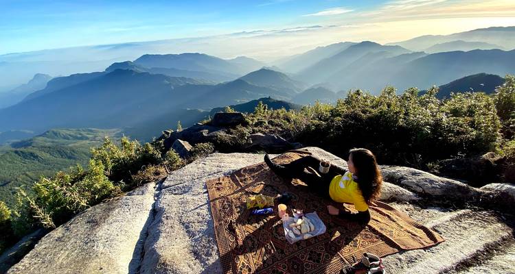Un voyageur solitaire se détend sur un tapis de pique-nique au sommet d'une falaise rocheuse, contemplant les couches de montagnes brumeuses sous la lumière chaude du lever de soleil.